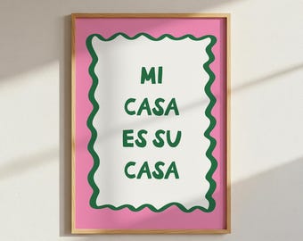 May include: A framed art print with the text "MI CASA ES SU CASA" in green on a white background, framed by a green wavy border, set against a pink backdrop. The frame is a light wood color.