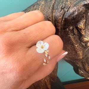 Puede incluir: Un anillo dorado con una flor blanca con centro dorado y un delicado diseño de hojas y cristales. El anillo se lleva en un dedo, mostrando sus intrincados detalles florales y su elegante estilo. El diseño del anillo incluye pequeños cristales brillantes.