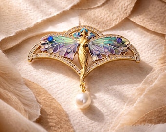 May include: A gold brooch in the shape of a fairy with outstretched wings, painted in shades of purple, green, and blue. The brooch is embellished with small, clear crystals and a dangling pearl. The brooch is set on a cream-coloured fabric.