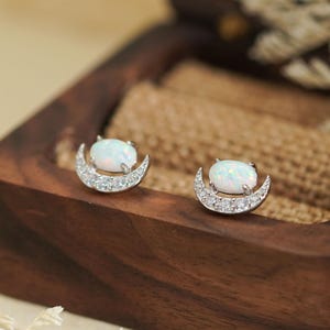 May include: A pair of silver-toned stud earrings featuring a crescent moon design. Each earring has a white opal stone at the center, surrounded by small, clear crystals. The earrings are displayed in a dark wooden jewelry box.