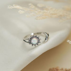 May include: A silver ring featuring a sun and moon design. The sun has a white opal center and detailed rays. The moon is a crescent shape. The ring is set against a white fabric background with dried plants.