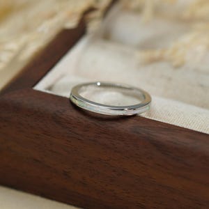 May include: A silver ring with a white band, displayed in a wooden jewellery box. The ring has a simple, polished design. The box is dark brown with a cream-coloured interior. The background is blurred, with hints of dried flowers. The ring is a classic piece of jewellery.