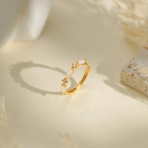 May include: A gold-toned ring featuring three marquise-shaped white stones arranged along the band, with small clear accent stones placed between them. The metal band is thin and polished, showcasing a delicate design with a bright, reflective finish.