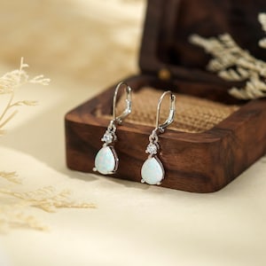 May include: A pair of silver-toned drop earrings with pear-shaped opal-like stones. Each earring features a small, round accent stone above the larger, teardrop-shaped opal. The earrings are displayed in an open, wooden jewelry box.