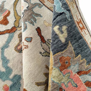 May include: A woven rug featuring a cream-colored base with a detailed floral and geometric pattern in shades of slate blue, burnt orange, sage green, and soft pink. The texture shows a dense pile with visible knots and a folded corner edge.