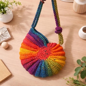 May include: A handmade, colorful crochet bag with a rainbow pattern. The bag features a semi-circular shape with a spiral design, showcasing shades of red, orange, yellow, green, blue, and purple. The strap is a twisted cord in coordinating colors.