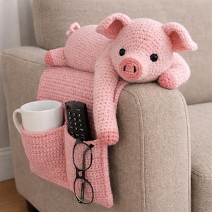 May include: A pink crocheted pig-shaped couch armrest organizer. The organizer has pockets for a white mug, remote control, and glasses. The pig is lying on the armrest. The organizer is made of pink yarn.