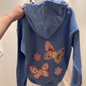 May include: A pale blue hoodie with butterfly and flower appliqués. The butterfly designs have floral and strawberry patterns. The hoodie is suspended from a white hanger.