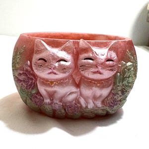 May include: A pink resin planter featuring two smiling cat figures. The cats are adorned with delicate collars and are surrounded by floral and foliage details. The planter has a rounded shape and a smooth finish, suitable for small plants or decorative use.