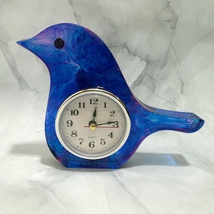 May include: A bird-shaped desk clock featuring a swirling blue and purple resin body. The clock face is circular with a silver-tone rim, black Arabic numerals, and black hour, minute, and second hands. The word Quartz is printed in black.