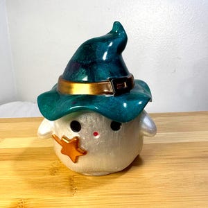 May include: A whimsical, pearlescent white figurine of a ghost wearing a teal witch hat with a gold band and buckle. A gold star adorns the ghost's cheek. The figurine has black eyes and a small red nose, perfect for Halloween decor.