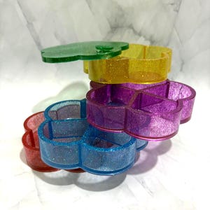 May include: A stack of four clover-shaped resin trinket dishes in translucent red, blue, purple, and yellow, each infused with fine glitter. The dishes feature a four-compartment interior design and are arranged in a vertical, overlapping formation.