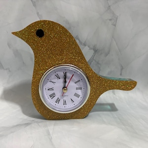May include: A bird-shaped desk clock covered in gold glitter. The clock face is white with black Roman numerals, a silver-toned bezel, and black hour and minute hands with a red second hand. A small black circle serves as the bird's eye.