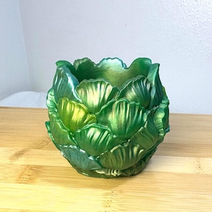 May include: A decorative, green artichoke-shaped planter. The planter has a metallic finish with shades of green and gold. The design features layered, textured petals, creating a unique and artistic look. This item is suitable for small plants or as a decorative piece.