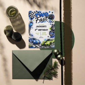May include: A rectangular birthday invitation card featuring the text TWO FAST and JACKSON'S 2ND BIRTHDAY, decorated with blue and green race cars, a checkered flag, and tire tracks. The card is paired with a dark green envelope on a neutral surface.