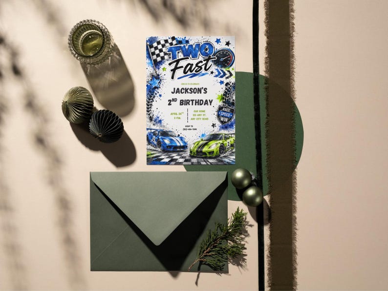 May include: A rectangular birthday invitation card featuring the text TWO FAST and JACKSON'S 2ND BIRTHDAY, decorated with blue and green race cars, a checkered flag, and tire tracks. The card is paired with a dark green envelope on a neutral surface.
