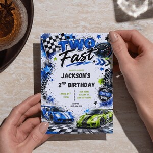May include: A rectangular birthday invitation card featuring a race car theme with blue and green sports cars, a checkered flag, and tire tracks. The text reads TWO FAST, JACKSON'S 2ND BIRTHDAY, APRIL 24TH, 3 P.M., OUR HOME, 123 ANY ST., ANY CITY 12345, and RSVP TO (555)-555-7890.