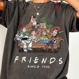 May include: A charcoal gray short-sleeve t-shirt featuring a graphic of Toy Story characters sitting on a couch with the text "F.R.I.E.N.D.S SINCE 1995" printed below. The characters include Buzz Lightyear, Woody, Jessie, Bullseye, Rex, Slinky Dog, and aliens.