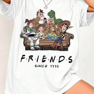 May include: A white t-shirt featuring a graphic print of various animated toy characters sitting on a brown couch. Below the couch, the text reads "F.R.I.E.N.D.S" with colorful dots between the letters, followed by the text "SINCE 1995" in black.