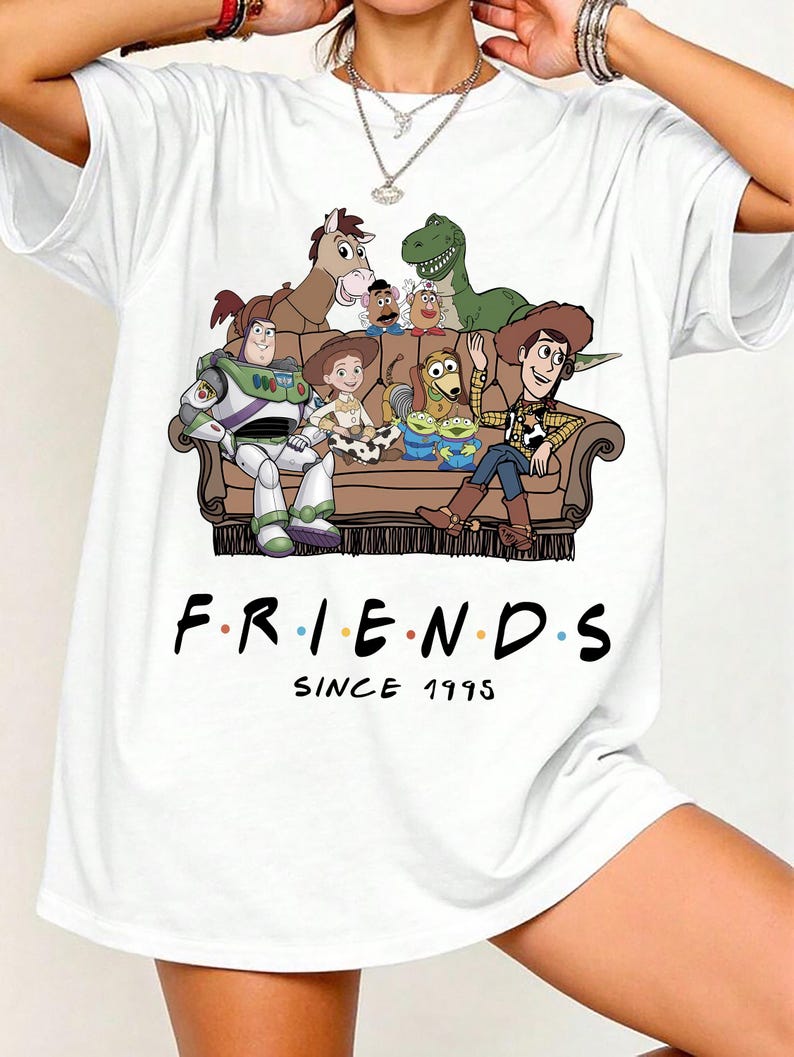May include: A white t-shirt featuring a graphic print of various animated toy characters sitting on a brown couch. Below the couch, the text reads "F.R.I.E.N.D.S" with colorful dots between the letters, followed by the text "SINCE 1995" in black.