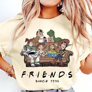 May include: A cream-colored t-shirt featuring a graphic of Toy Story characters sitting on a brown couch. The characters include Buzz Lightyear, Woody, Jessie, Rex, Bullseye, Slinky Dog, Mr. Potato Head, and Aliens. Below the couch is the text FRIENDS SINCE 1995.