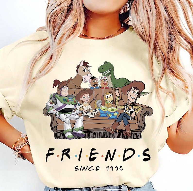 May include: A cream-colored t-shirt featuring a graphic of Toy Story characters sitting on a brown couch. The characters include Buzz Lightyear, Woody, Jessie, Rex, Bullseye, Slinky Dog, Mr. Potato Head, and Aliens. Below the couch is the text FRIENDS SINCE 1995.