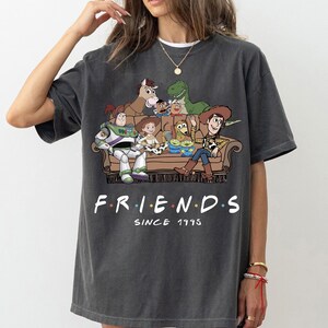 May include: A charcoal gray short-sleeve t-shirt featuring a graphic print of various animated toy characters sitting on a brown sofa. Below the illustration, the text reads "F.R.I.E.N.D.S" with colorful dots between letters and "SINCE 1995" underneath.