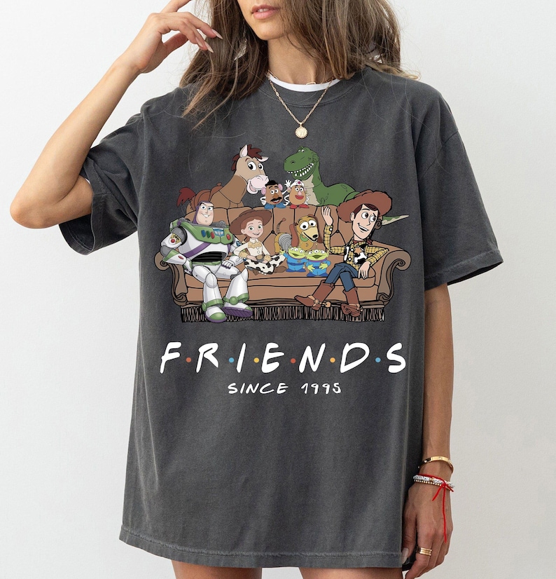 May include: A charcoal gray short-sleeve t-shirt featuring a graphic print of various animated toy characters sitting on a brown sofa. Below the illustration, the text reads "F.R.I.E.N.D.S" with colorful dots between letters and "SINCE 1995" underneath.