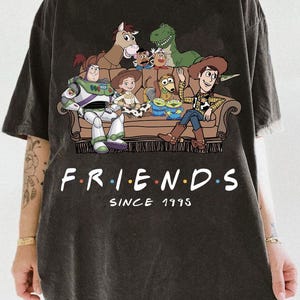 May include: A charcoal gray t-shirt featuring a graphic of Toy Story characters sitting on a brown couch. The text "F.R.I.E.N.D.S" is printed in white with colorful dots between letters, followed by the text "SINCE 1995" in a white serif font.