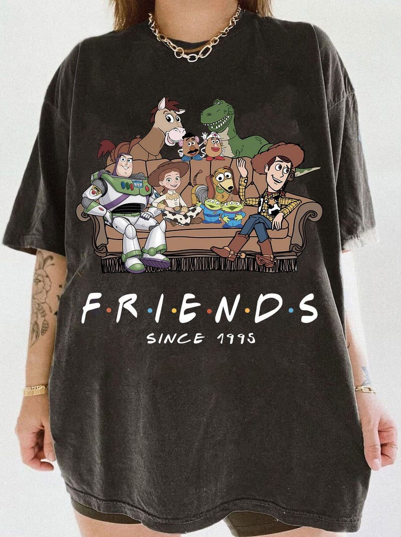May include: A charcoal gray t-shirt featuring a graphic of Toy Story characters sitting on a brown couch. The text "F.R.I.E.N.D.S" is printed in white with colorful dots between letters, followed by the text "SINCE 1995" in a white serif font.