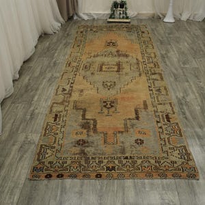 May include: A long rectangular area rug featuring a central diamond medallion pattern in shades of muted orange, gray, and brown. The rug displays intricate geometric borders and tribal-inspired motifs throughout its weathered, low-pile surface.