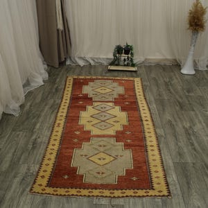 May include: A rectangular area rug featuring a deep terracotta red background with three large, centred geometric medallions in light grey and cream. The border is a cream colour with a repeating dark brown diamond pattern along the edges.