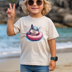 May include: A beige t-shirt with a cartoon shark in a pink donut float. The shark is blue with white teeth and red eyes, surrounded by water splashes. The t-shirt is worn by a child. The image is set on a beach.