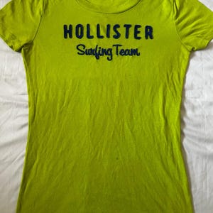 May include: A lime green short-sleeved t-shirt featuring the dark blue embroidered text HOLLISTER Surfing Team across the chest. The shirt has a crew neckline and a small interior brand label at the neck indicating a size small.