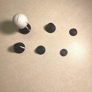 Puede incluir: Una pelota de golf blanca está equilibrada sobre un marcador de pelota de golf negro, rodeada de varios otros marcadores negros. Los marcadores tienen un diseño circular. Los artículos están sobre una superficie beige claro.