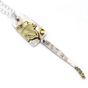 May include: A silver and gold necklace with a pendant featuring a textured design and a small yellow gemstone. The pendant is shaped like a leaf and has a textured design.