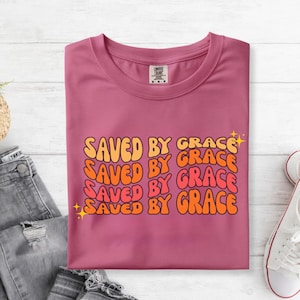 May include: A mauve t-shirt with the text "SAVED BY GRACE" in orange and yellow retro font. The shirt is folded, with a pair of jeans and white sneakers visible in the background.