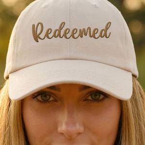 Puede incluir: Gorra de béisbol beige con la palabra "Redeemed" bordada en una fuente cursiva. La gorra tiene una visera curva y un diseño clásico de seis paneles. El texto es de color marrón cálido.