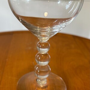 May include: A clear glass champagne coupe with a wide, shallow bowl and a stem composed of stacked spheres. The base is a wide, flat circle. The glass is designed for serving sparkling wine or cocktails.