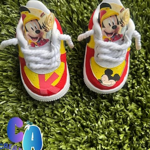 May include: Pair of custom baby trainers in red, yellow, and white. The shoes feature a Mickey Mouse design on the side and a Mickey Mouse image on the tongue. White laces and red and white soles complete the design.