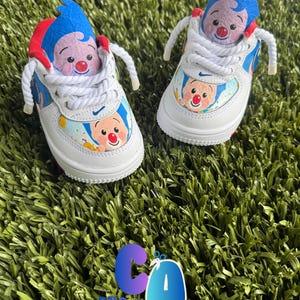 May include: A pair of white sneakers with custom artwork featuring a clown design. The shoes have blue, red, and white accents, with a clown figure on the tongue and sides. The laces are white, and the shoes are set on a green surface.