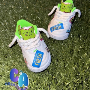 Pode incluir: Sapatilhas brancas para bebé com detalhes verdes e rosa. As sapatilhas têm atacadores brancos e uma etiqueta azul que diz "California Fresh". A língua do sapato tem texto verde. Os sapatos estão numa superfície verde.