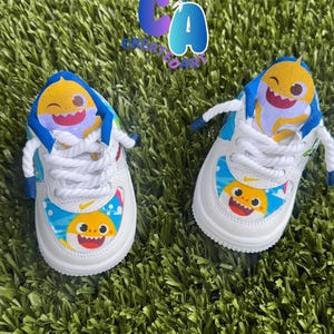 May include: White baby trainers with a Baby Shark theme. The shoes have a yellow shark graphic on the toe and tongue, with blue and white details. White laces and a green background complete the look.
