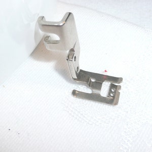 May include: A silver sewing machine foot with a metal attachment. The foot is designed for sewing straight seams.