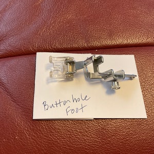 Peut inclure: Un pied de boutonni&egrave;re pour machine &agrave; coudre en argent avec un accessoire en plastique transparent. Le pied est &eacute;tiquet&eacute; "Buttonhole Foot" sur un morceau de papier blanc.