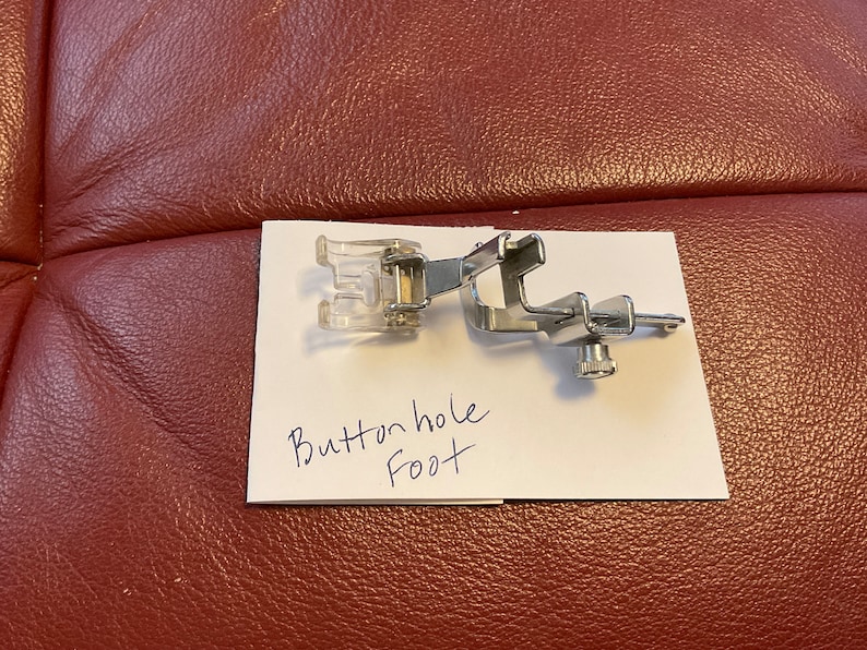 Peut inclure: Un pied de boutonni&egrave;re pour machine &agrave; coudre en argent avec un accessoire en plastique transparent. Le pied est &eacute;tiquet&eacute; "Buttonhole Foot" sur un morceau de papier blanc.