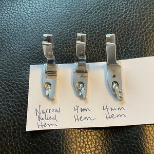 Peut inclure: Trois pieds-de-biche en argent pour machine &agrave; coudre pour cr&eacute;er des ourlets roul&eacute;s. Les pieds sont &eacute;tiquet&eacute;s "Ourlet roul&eacute; &eacute;troit", "Ourlet de 4 mm" et "Ourlet de 4 mm".