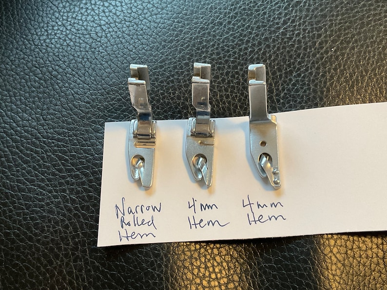 Peut inclure: Trois pieds-de-biche en argent pour machine &agrave; coudre pour cr&eacute;er des ourlets roul&eacute;s. Les pieds sont &eacute;tiquet&eacute;s "Ourlet roul&eacute; &eacute;troit", "Ourlet de 4 mm" et "Ourlet de 4 mm".
