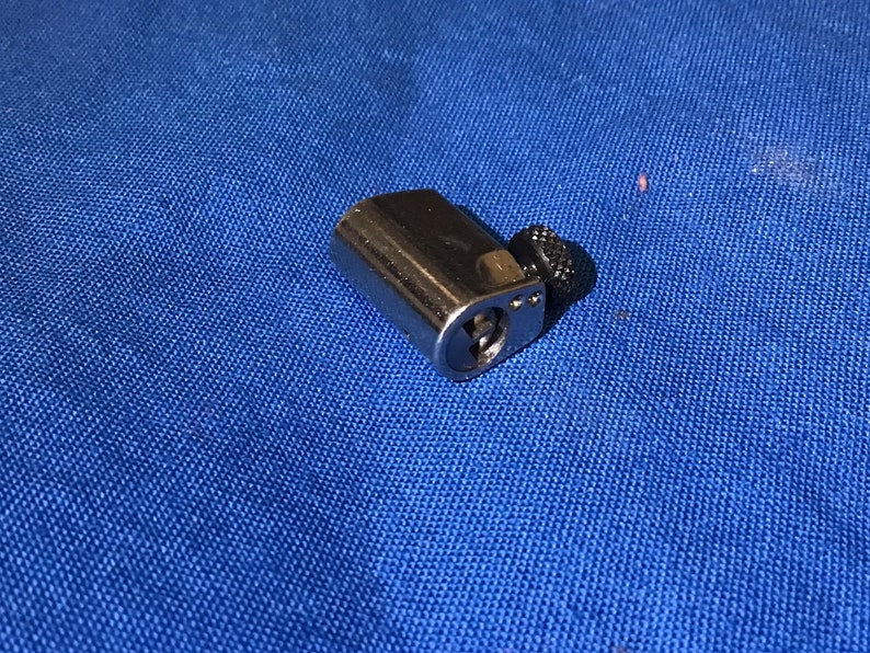 Może przedstawiać: Srebrny cylinder zamka z czarnym pokrętłem. Cylinder ma dziurkę od klucza i mały metalowy kołek.