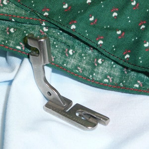 May include: A silver sewing machine foot with a curved metal arm and a flat base. The foot is labeled "TWINCO MADE IN JAPAN".